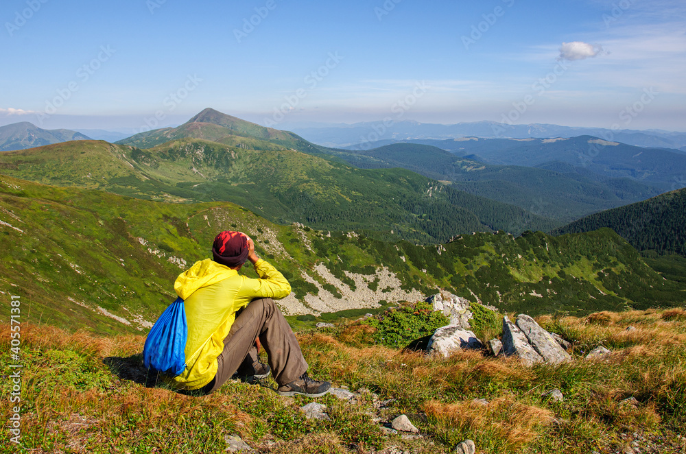 Fototapeta premium A man in a yellow jacket sits on a mountainside and looks at the goverla, the highest peak of the Carpathian mountains in Ukraine