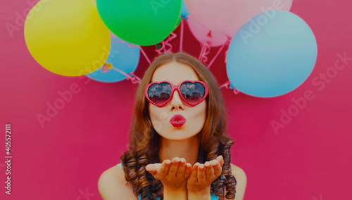 Portrait of beautiful woman blowing her red lips sending air kiss with colorful balloons on a pink background