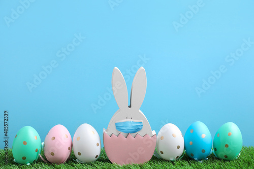 COVID-19 pandemic. Easter bunny figure in protective mask and dyed eggs on green grass against light blue background