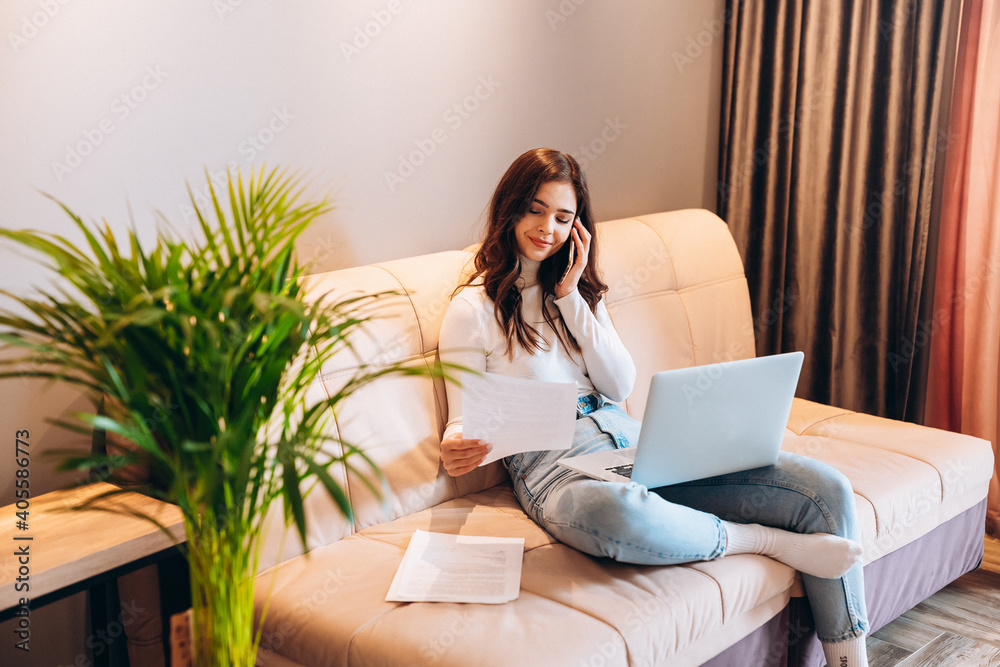 Freelance student young female using laptop while sitting at home. Young woman sitting on sofa and working online on laptop. Pretty female talking on phone while studying working Freelance student young female using laptop while sitting at home. Young woman sitting on sofa and working online on laptop. Pretty female talking on phone while studying working