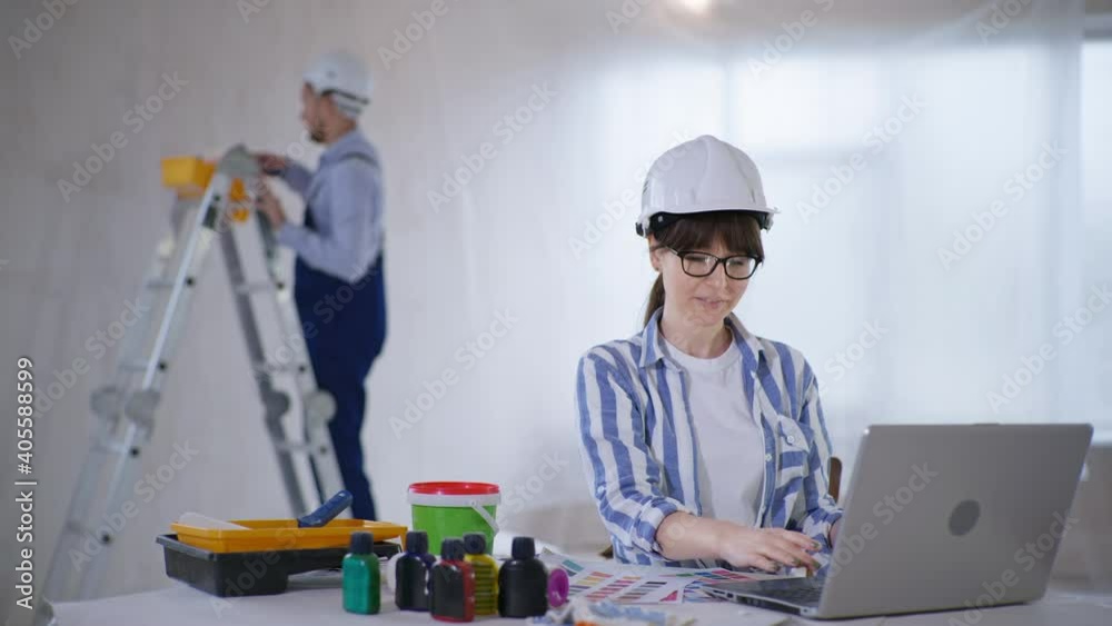 young beautiful woman working as an interior designer chooses color of paint for walls, while sitting at computer background of male painter doing repairs in an apartment young beautiful woman working as an interior designer chooses color of paint for walls, while sitting at computer background of male painter doing repairs in an apartment