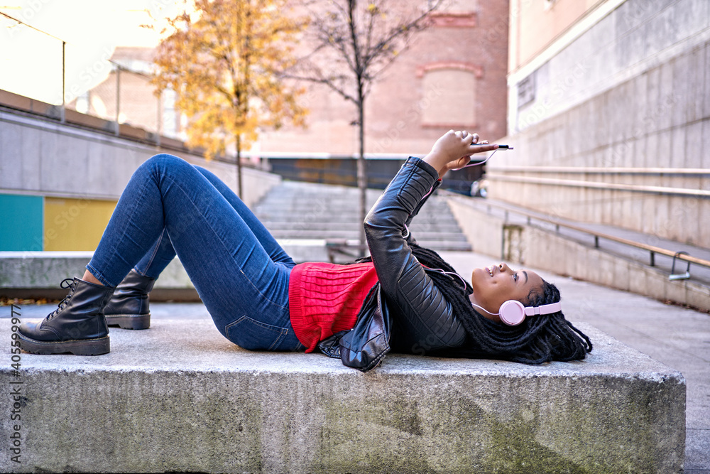 Portrait of a smiling African-American woman with a cell phone is chatting or listening to music while lying on a city bench. The hair is braided into dreadlocks.