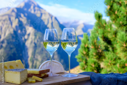 Tasty cheese and wine from Savoy region in France, beaufort, abondance, emmental, tomme and reblochon de savoie cheeses and glass of white wine served outdoor with Alpine mountains peaks on background photo