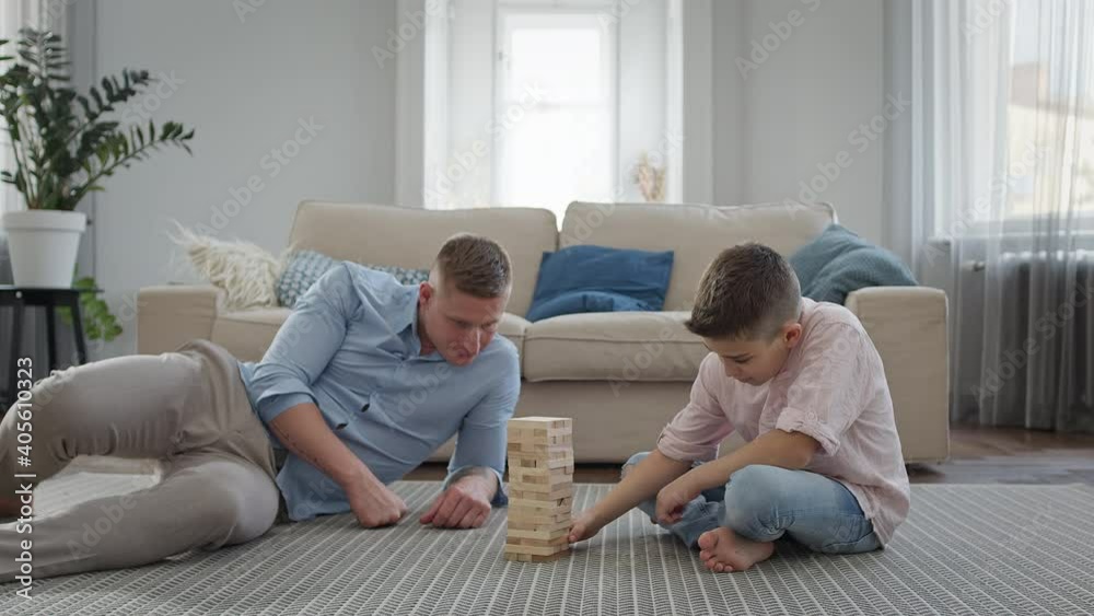 Father And Son Sitting On The Floor In The Apartment. They Play Game Of Assembling Pyramid Of Wooden Bars. Father And Son Sitting On The Floor In The Apartment. They Play Game Of Assembling Pyramid Of Wooden Bars.
