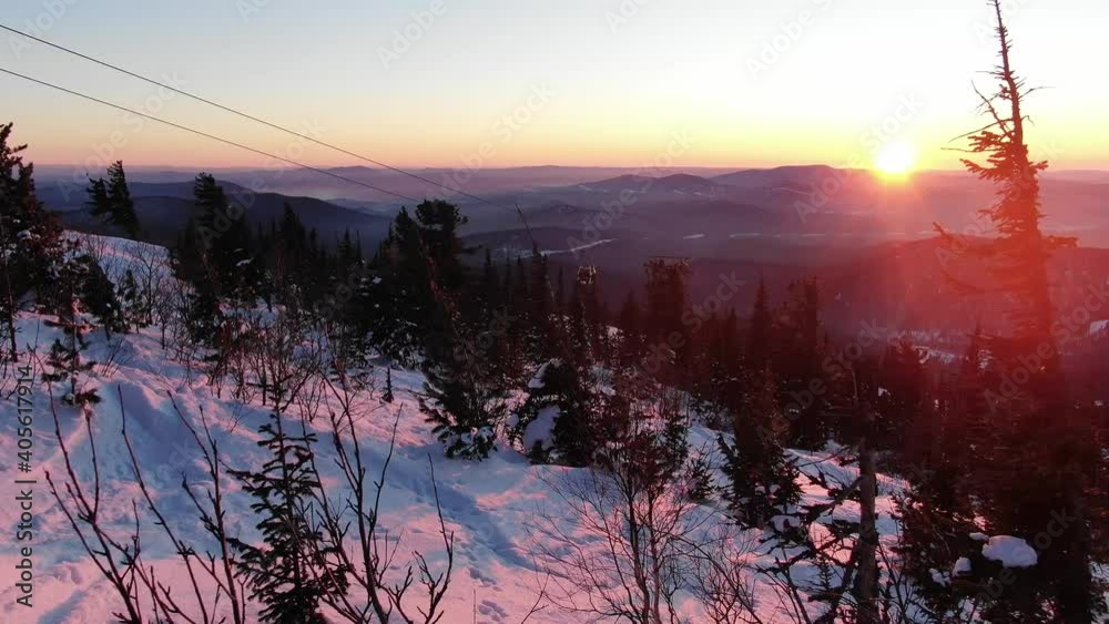 Flying through snow-covered treetops and over ski lift cabins at sunset against background of mountains in rays of departing sun. Outdoor activities at ski resort during vacation.