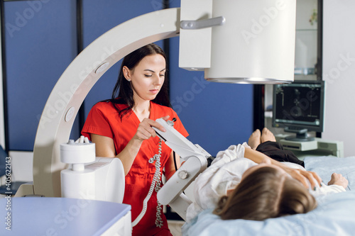 Young attractive focused woman doctor in red uniform, providing lithotripsy procedure for her lying female patient with modern ultrasonic lithotriptor to break up stones photo