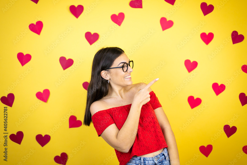 Young beautiful woman over yellow background with red hearts smiling and pointing with hand and finger to the side Young beautiful woman over yellow background with red hearts smiling and pointing with hand and finger to the side