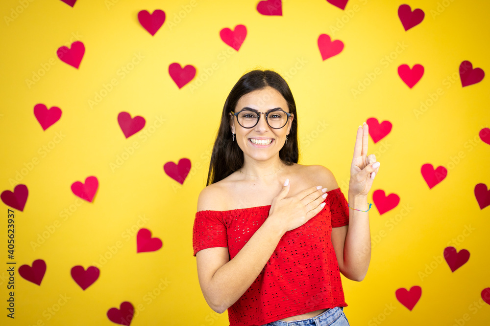 Young beautiful woman over yellow background with red hearts smiling swearing with hand on chest and fingers up, making a loyalty promise oath Young beautiful woman over yellow background with red hearts smiling swearing with hand on chest and fingers up, making a loyalty promise oath