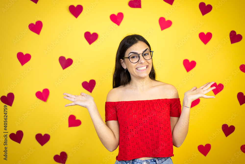 Young beautiful woman over yellow background with red hearts clueless and confused expression with arms and hands raised Young beautiful woman over yellow background with red hearts clueless and confused expression with arms and hands raised