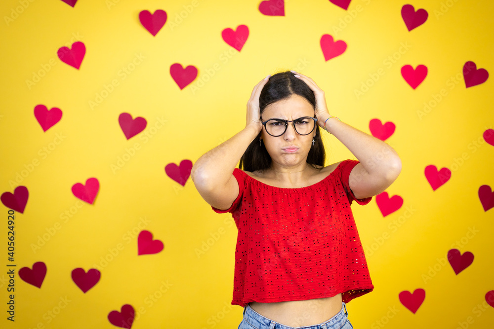Young beautiful woman over yellow background with red hearts thinking looking tired and bored with hands on head Young beautiful woman over yellow background with red hearts thinking looking tired and bored with hands on head