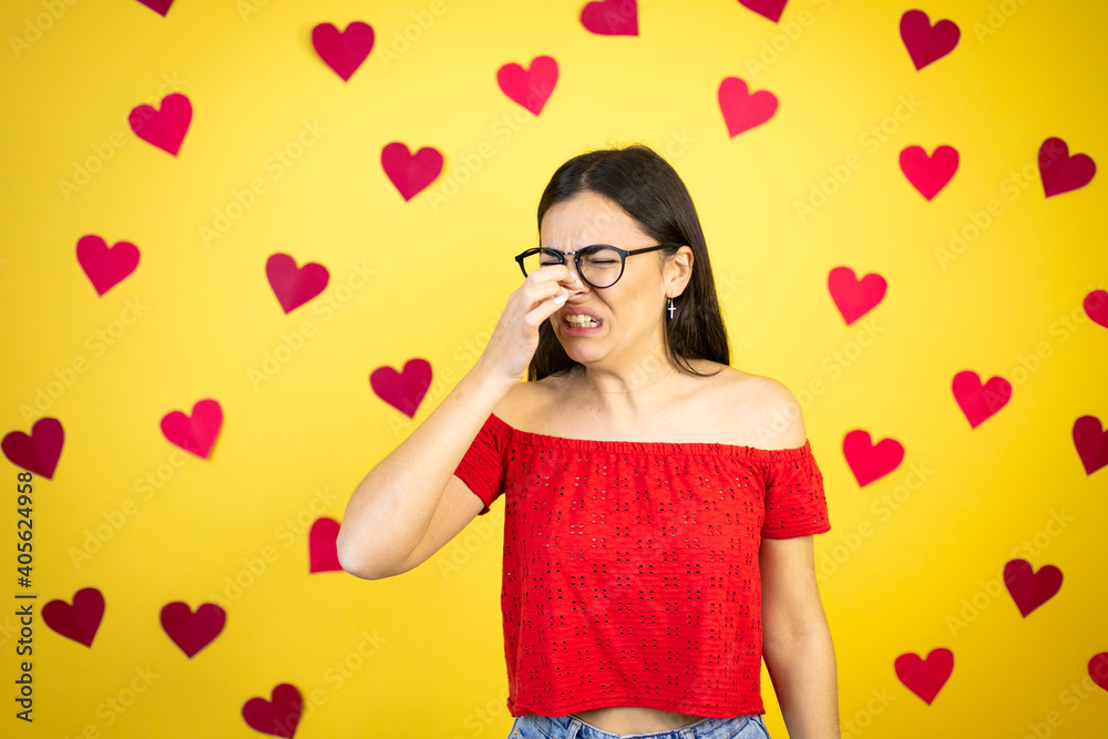 Young beautiful woman over yellow background with red hearts smelling something stinky and disgusting, intolerable smell, holding breath with fingers on nose Young beautiful woman over yellow background with red hearts smelling something stinky and disgusting, intolerable smell, holding breath with fingers on nose