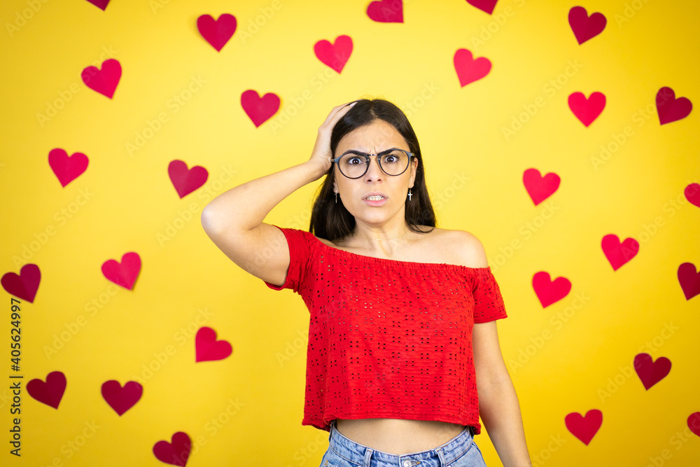 Young beautiful woman over yellow background with red hearts putting one hand on her head smiling like she had forgotten something