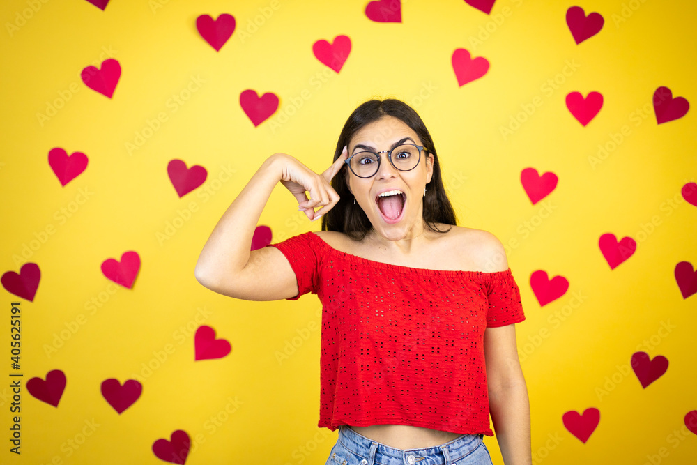 Young beautiful woman over yellow background with red hearts smiling and thinking with her fingers on her head that she has an idea. Young beautiful woman over yellow background with red hearts smiling and thinking with her fingers on her head that she has an idea.