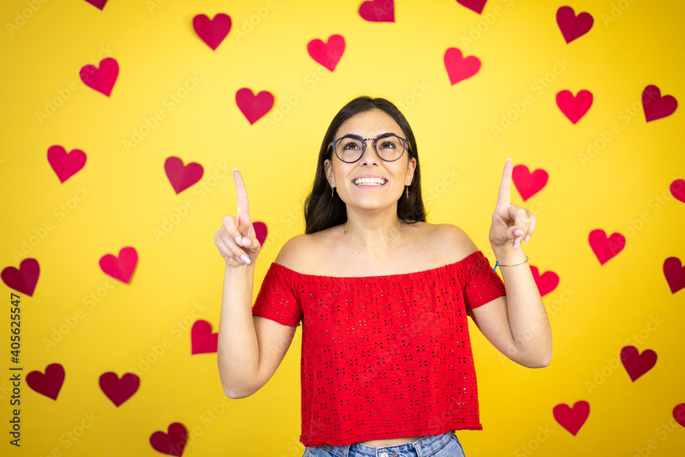 Young beautiful woman over yellow background with red hearts smiling, looking at the camera and pointing up with fingers and raised arms Young beautiful woman over yellow background with red hearts smiling, looking at the camera and pointing up with fingers and raised arms