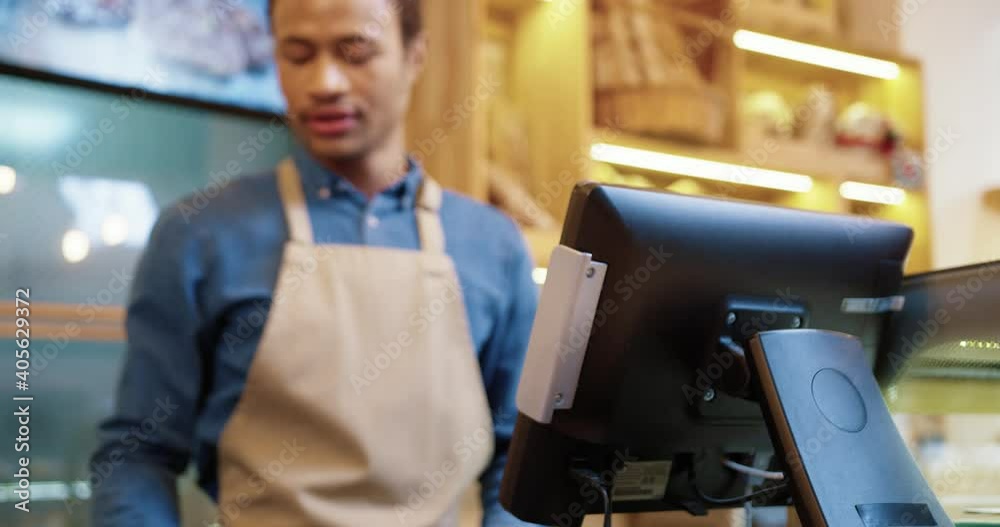 Young African-American salesman in bakery is counting on contactless customer with bank terminal. Concept of cashless payment for online banking services by smartphone. Young African-American salesman in bakery is counting on contactless customer with bank terminal. Concept of cashless payment for online banking services by smartphone.