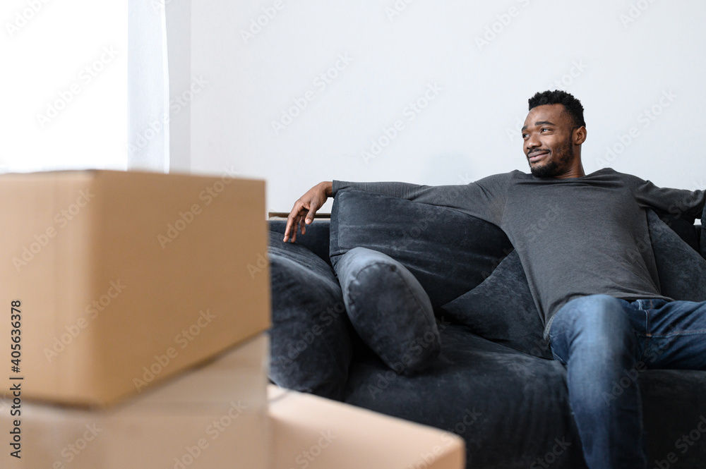 Smiling single male sitting on the sofa around cardboard boxes, moving at new apartment. Successful african american man buy a real estate by his own, achieved a goal. Relocation day concept