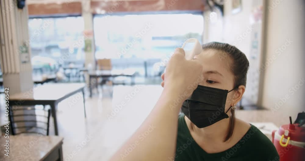 close up shot of staff using infrared thermometer checker on young Asian woman wearing facial protective mask customer's forehead before enter, waiting for Temperature checkpoints in restaurant,