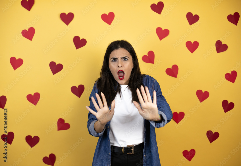 Young caucasian woman over yellow background with red hearts afraid and terrified with fear expression stop gesture with hands, shouting in shock. Panic concept. Young caucasian woman over yellow background with red hearts afraid and terrified with fear expression stop gesture with hands, shouting in shock. Panic concept.