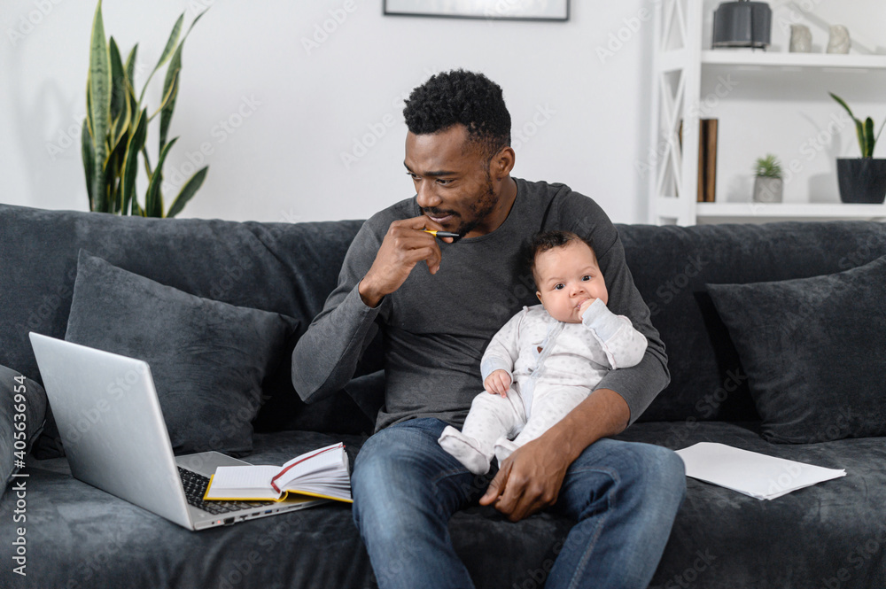 A thoughtful freelancer, student dad with an infant sit on the couch, he works from home. A young African-American father using laptop for remote work, while looking after his cute baby daughter A thoughtful freelancer, student dad with an infant sit on the couch, he works from home. A young African-American father using laptop for remote work, while looking after his cute baby daughter