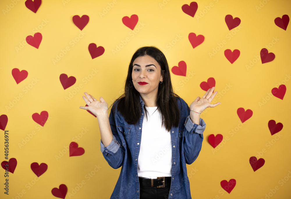 Young caucasian woman over yellow background with red hearts clueless and confused expression with arms and hands raised