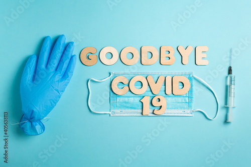 Words GOODBYE COVID 19 from wooden letters and syringe with vaccine on a blue background, inflated medical glove waving bye-bye and facemask. Vaccination and the end of epidemic concept.