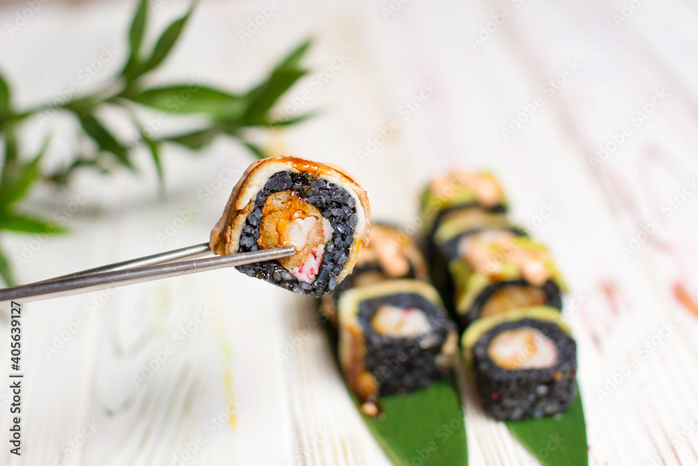 Close up of Sushi Roll piece with black rice and unagi eel on top in metal chopsticks with blurred background. Sushi Roll with crunchy tempura shrimp, bamboo leaves and plant on background. Copy space Close up of Sushi Roll piece with black rice and unagi eel on top in metal chopsticks with blurred background. Sushi Roll with crunchy tempura shrimp, bamboo leaves and plant on background. Copy space