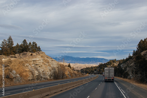 A road landscape on an autumn day, a highway on the sides of which there are sheer rocks with large stones, among which trees grow, trucks are driving, a beautiful sky, mountains in the distance. © Liudmila