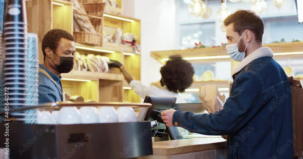 Young Caucasian customer in protective medical mask buys bread in bakery and pays by credit card. Concept of physical shopping with social distance during epidemic.