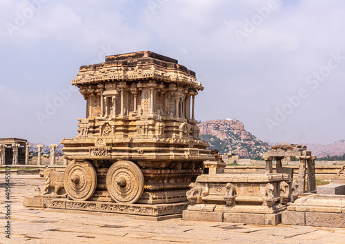Hampi, Karnataka, India - November 5, 2013: Vijaya Vitthala Temple. Beige stone chariot statue in center of complex under light blue sky. Sculptures and mountain on horizon.