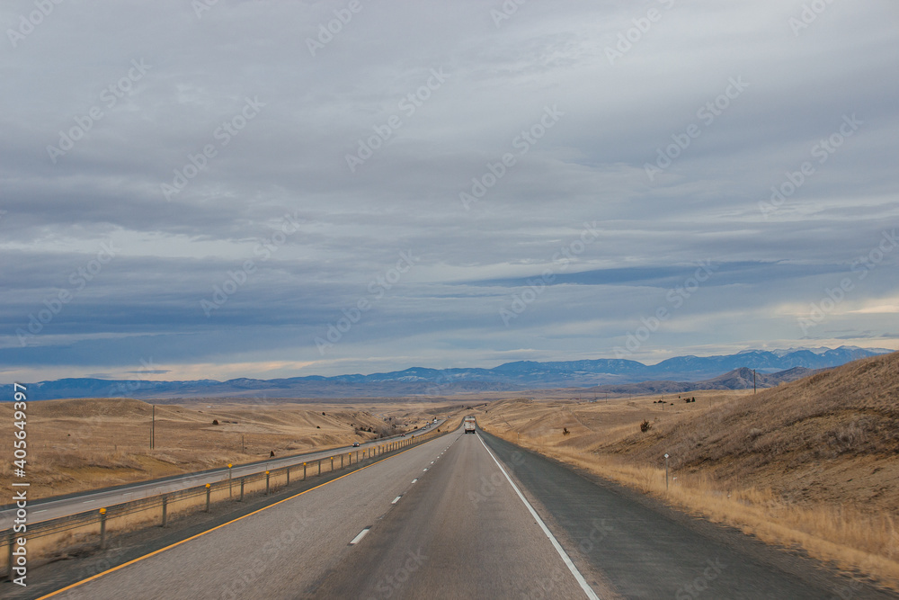 Asphalt road among steppes with yellow dry grass, blue mountains on the horizon, beautiful blue-gray autumn sky. Montana, USA, 11-23-2019