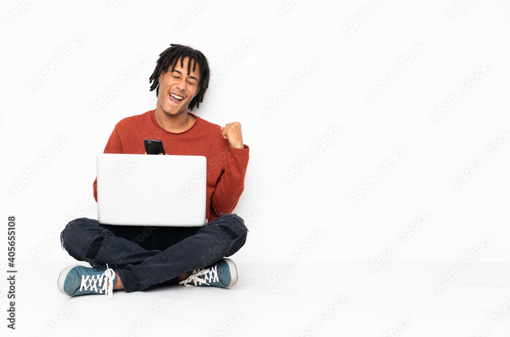 Young african american man sitting on the floor and working with his laptop with phone in victory position Young african american man sitting on the floor and working with his laptop with phone in victory position