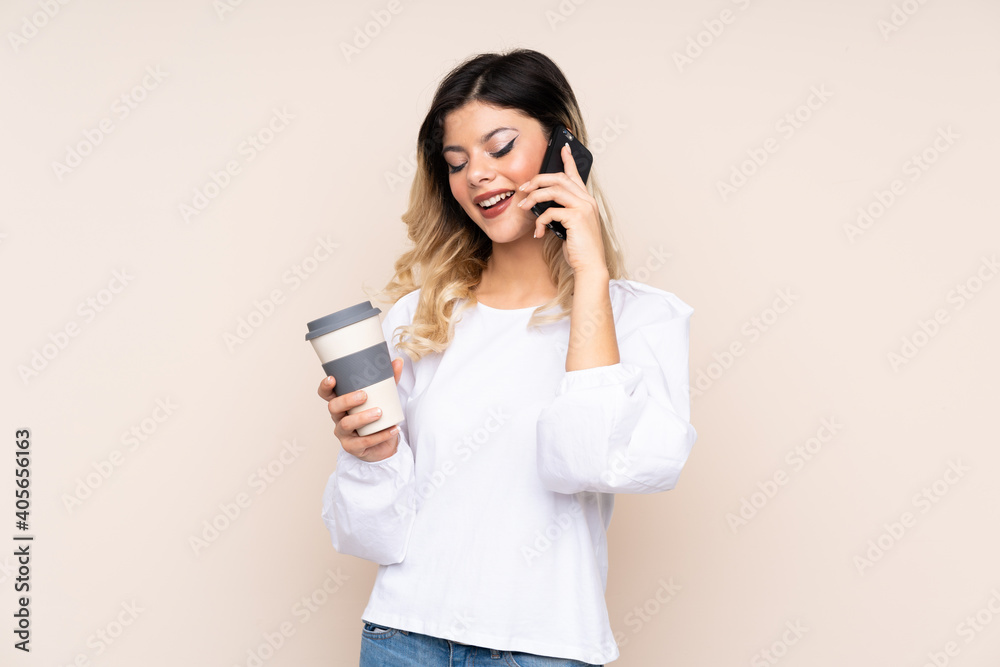 Teenager girl isolated on beige background holding coffee to take away and a mobile