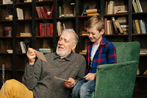 Fotografia Senior bearded gray-haired grandfather with his grandson enjoy memories watching family photo album sitting on armchair and floor at home in living room with an authentic aristocratic interior