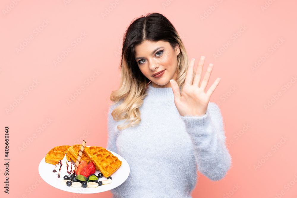 Teenager girl holding waffles on isolated pink background saluting with hand with happy expression