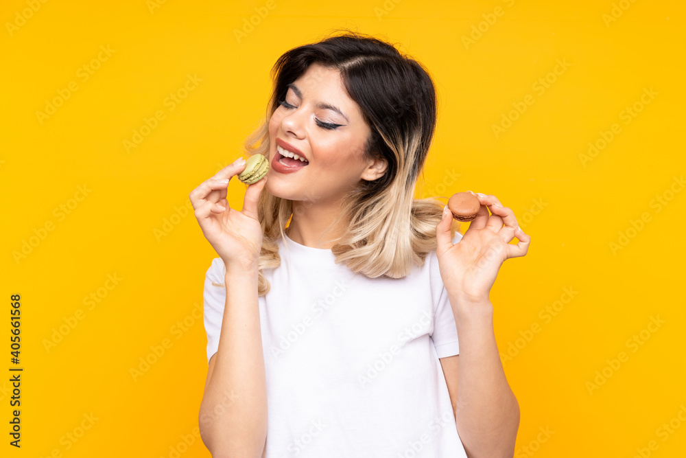 Teenager girl isolated on yellow background holding colorful French macarons and eating it Teenager girl isolated on yellow background holding colorful French macarons and eating it