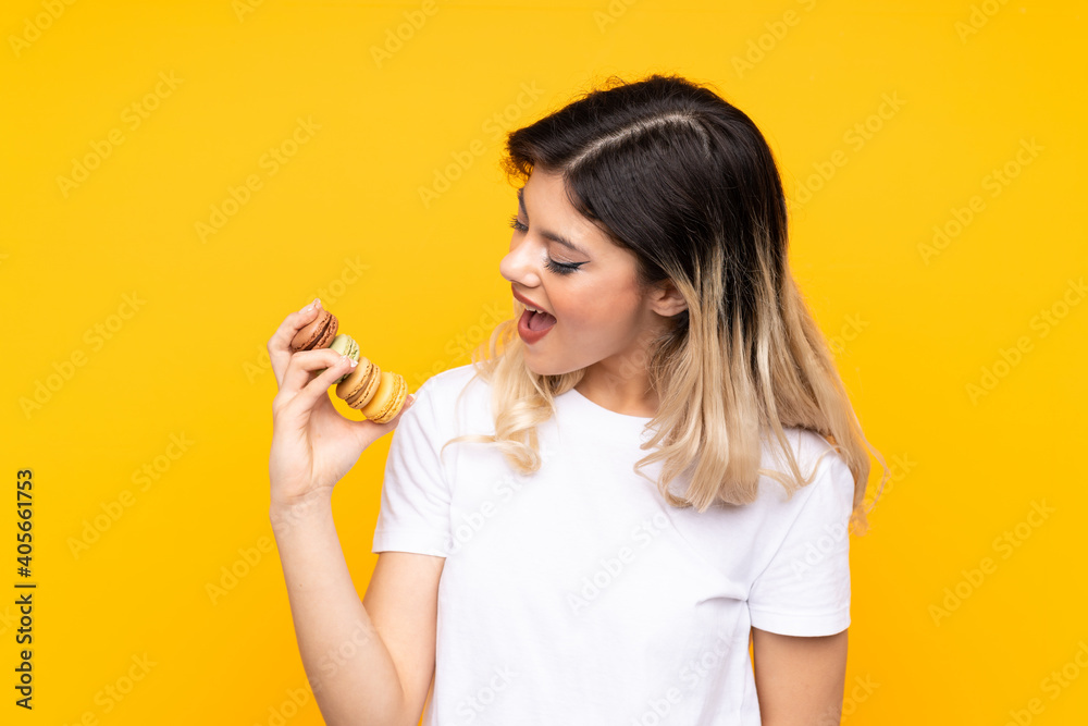 Teenager girl isolated on yellow background holding colorful French macarons and celebrating a victory Teenager girl isolated on yellow background holding colorful French macarons and celebrating a victory