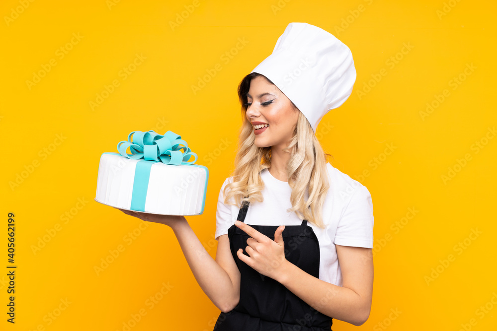 Teenager girl pastry holding a big cake isolated on yellow background pointing finger to the side Teenager girl pastry holding a big cake isolated on yellow background pointing finger to the side