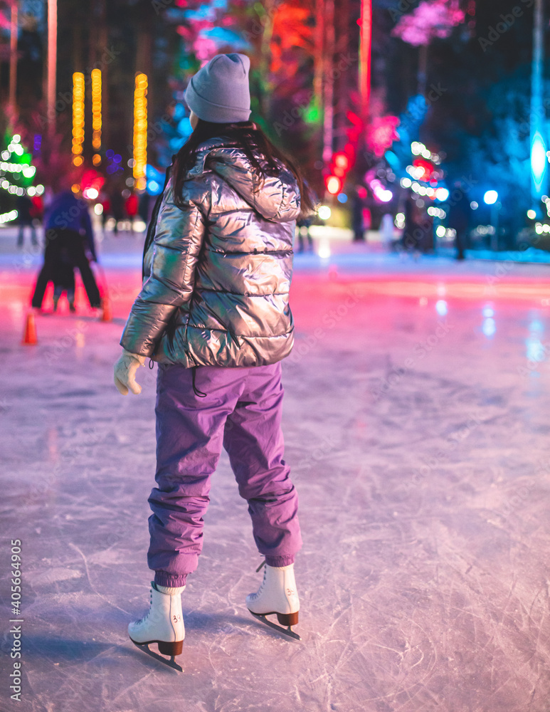 Naklejka premium Girl ice skating on the ice rink arena with happy people in the background, concept of ice skating in winter, winter activities, holiday christmas time, with new year decoration and illumination