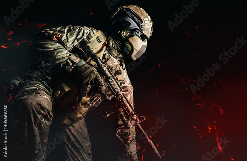 Obraz na plátně Photo of a fully equipped soldier in armored vest, helmet, face glasses and protection kneeling and attacking with rifle on dark background with ashes flying