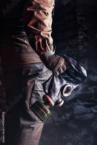 Photo of stalker hand in jacket and gloves holding old solviet rubber gas mask on destructed bricks background.