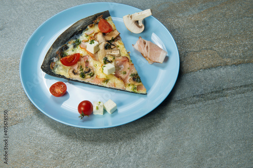 Slice of black dough pizza with ingredients on blue plate on stone anthracite background. Piece of pizza with cherry tomatoes, champion mushrooms, bacon, feta cheese served with basil pesto sauce