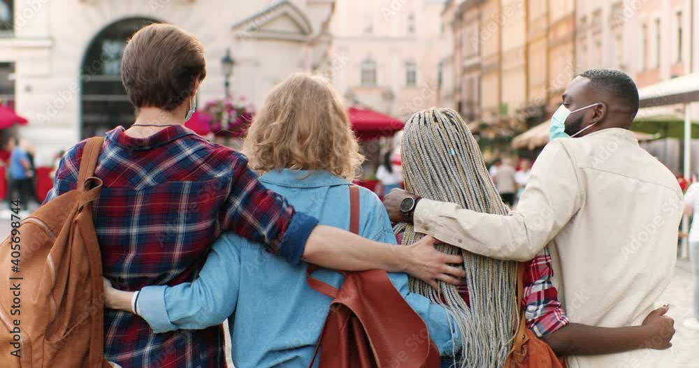 Rear of mixed-races males and females friends tourists in medical masks walking in city in hugs and having fun. Multi ethnic young men and women embracing, strolling in town and talking. Back view. Rear of mixed-races males and females friends tourists in medical masks walking in city in hugs and having fun. Multi ethnic young men and women embracing, strolling in town and talking. Back view.