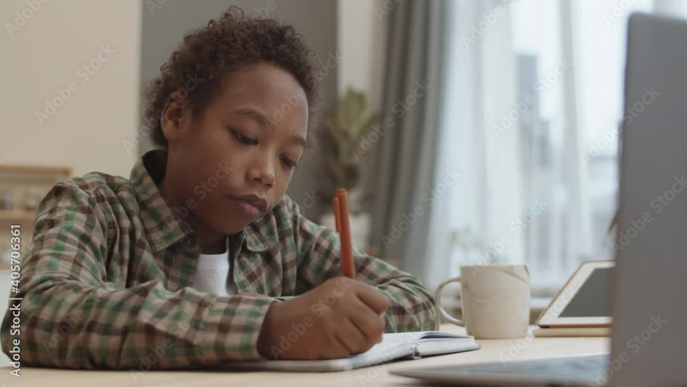 Chest-up of African ten-year-old male student sitting by desk at home, writing in notebook, looking at laptop computer in front of him, doing homework alone Chest-up of African ten-year-old male student sitting by desk at home, writing in notebook, looking at laptop computer in front of him, doing homework alone