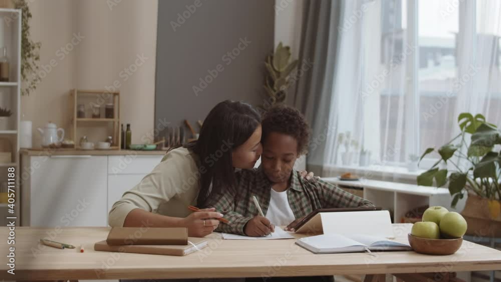 Chest-up of loving Mixed-Race mother kissing on cheek ten-year-old African son sitting by desk at home doing homework, writing on paper