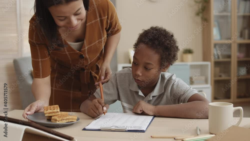 Chest up of ten-year-old African boy sitting by desk at home, doing homework, writing on paper, caring Mixed-Race mother bringing him plate of waffles and leaving