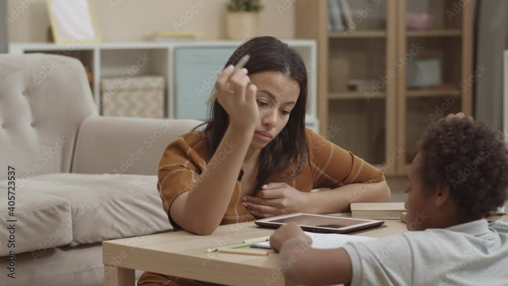Over shoulder chest-up of young irritated Mixed-Race woman sitting on floor at table in living room, lecturing her curly-haired child, crossing mistakes in his homework