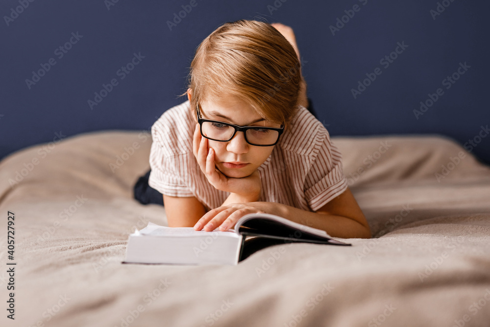 A 10-year-old boy with glasses is lying on the bed reading a big book A 10-year-old boy with glasses is lying on the bed reading a big book