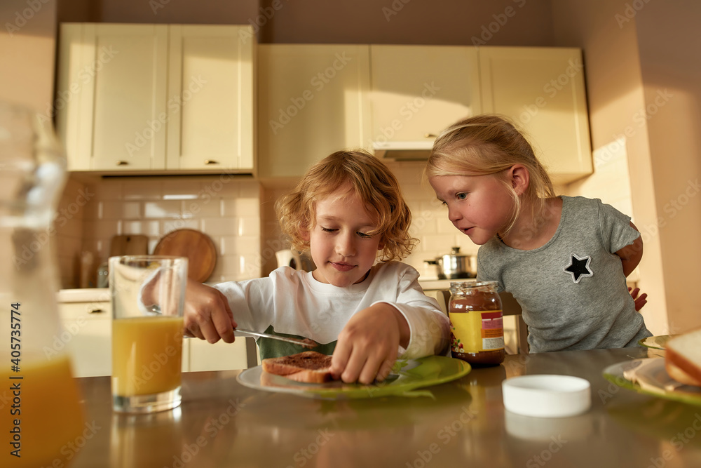 Portrait of cute little girl watching her brother spreading chocolate butter on toasted bread while preparing lunch or breakfast for his sister and himself in the kitchen Portrait of cute little girl watching her brother spreading chocolate butter on toasted bread while preparing lunch or breakfast for his sister and himself in the kitchen