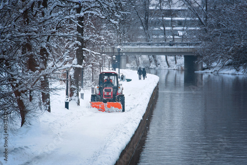 Canvas Print Tractor with snow plow and rotating brush sweeping snow from foopath on embankment in park