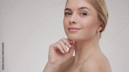 Beauty face. Young woman touching healthy skin portrait. Beautiful smiling girl with fresh glowing hydrated facial skin and natural makeup on light background at studio. Skin care concept. Slow motion